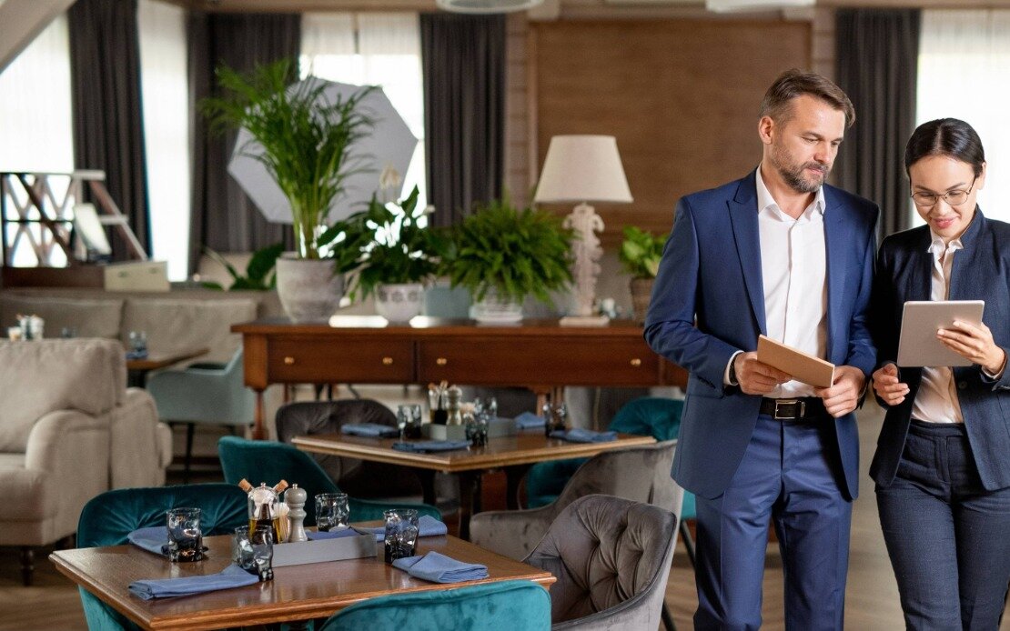 Ein Mann und eine Frau im Anzug stehen in einem modern eingerichtetem Restaurant und schauen auf einen kleinen Bildschirm