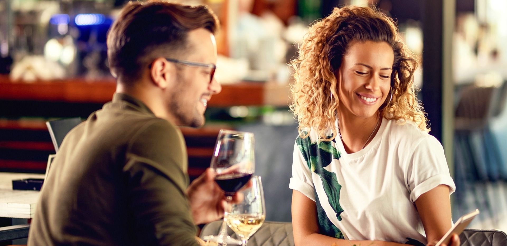 Eine Frau und ein Mann sitzen mit einem Glas Wein in einem Restaurant und bestellen lächelnd etwas über ihr Smartphone